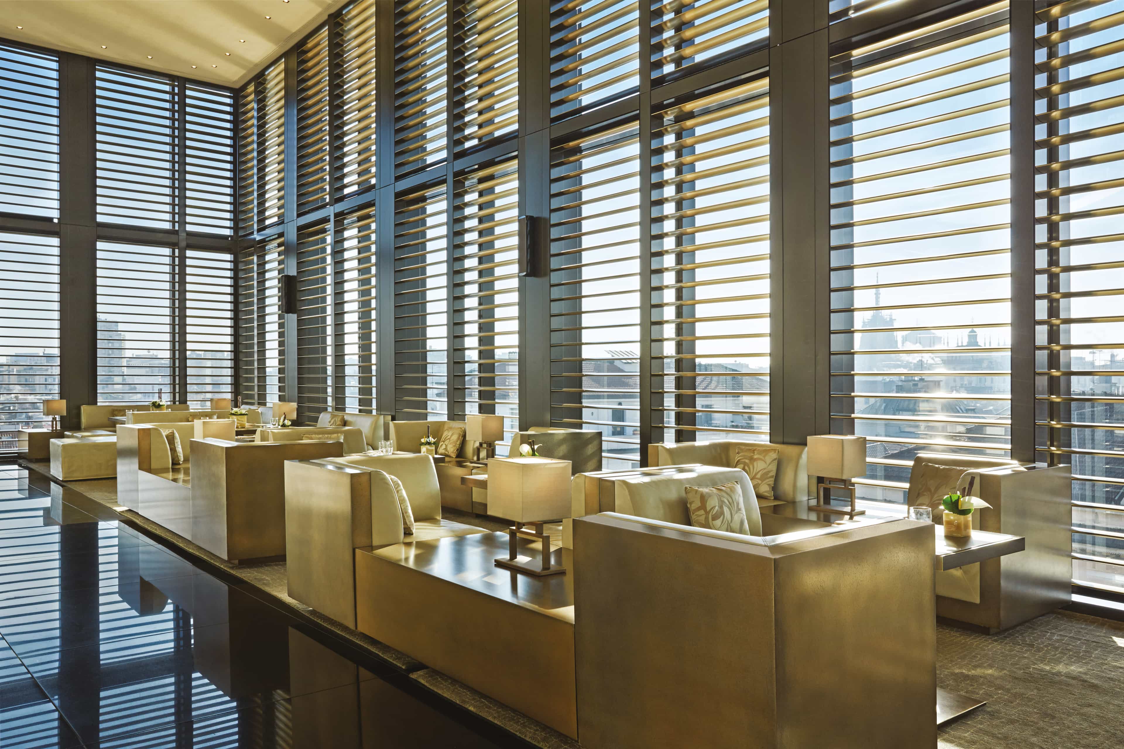 A hotel bar area with gold, plus seating and floor-to-ceiling windows with shutters showing cityscape