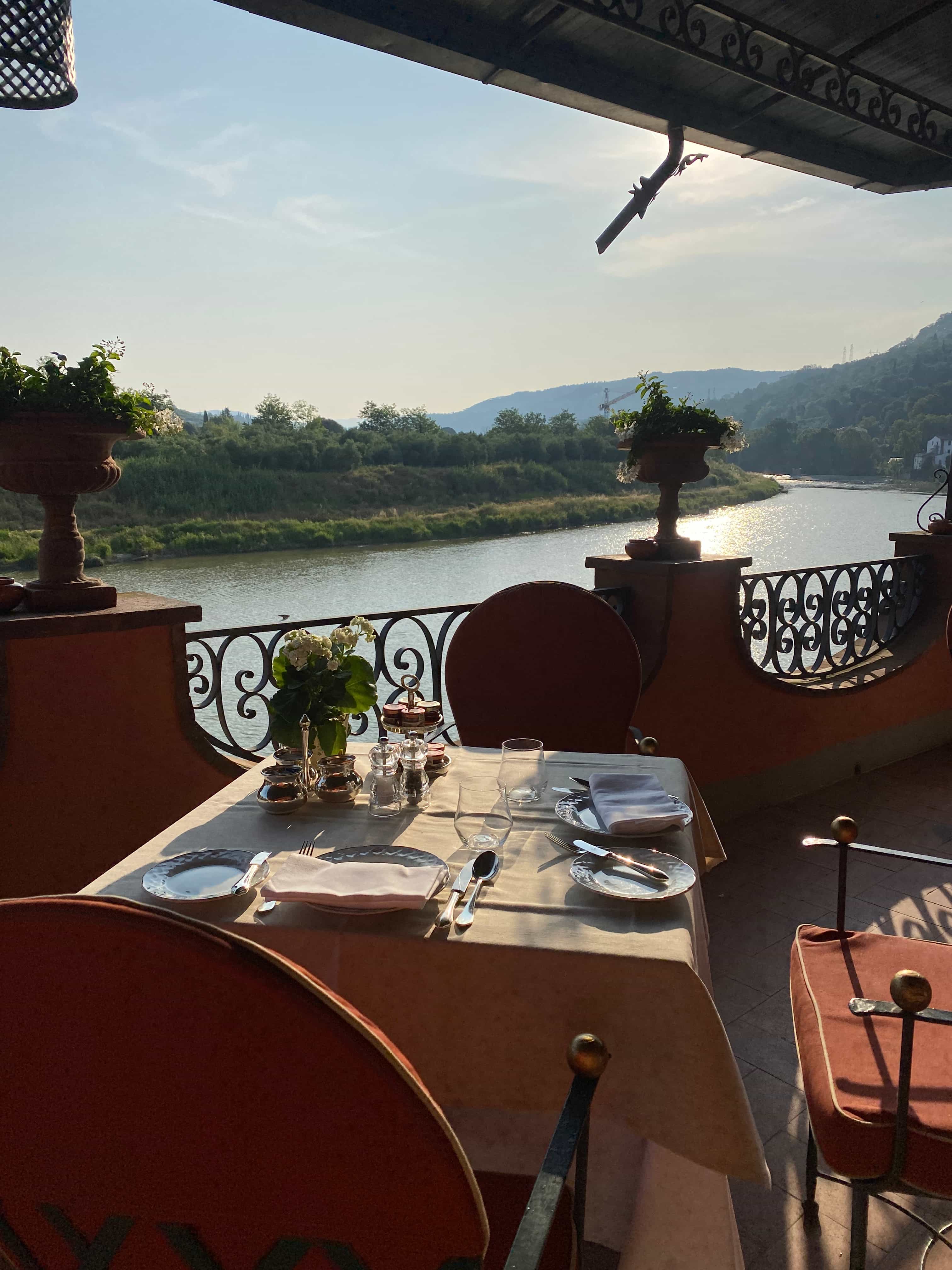 An outdoor restaurant table set for two on a balcony overlooking a beautiful river on a sunny day