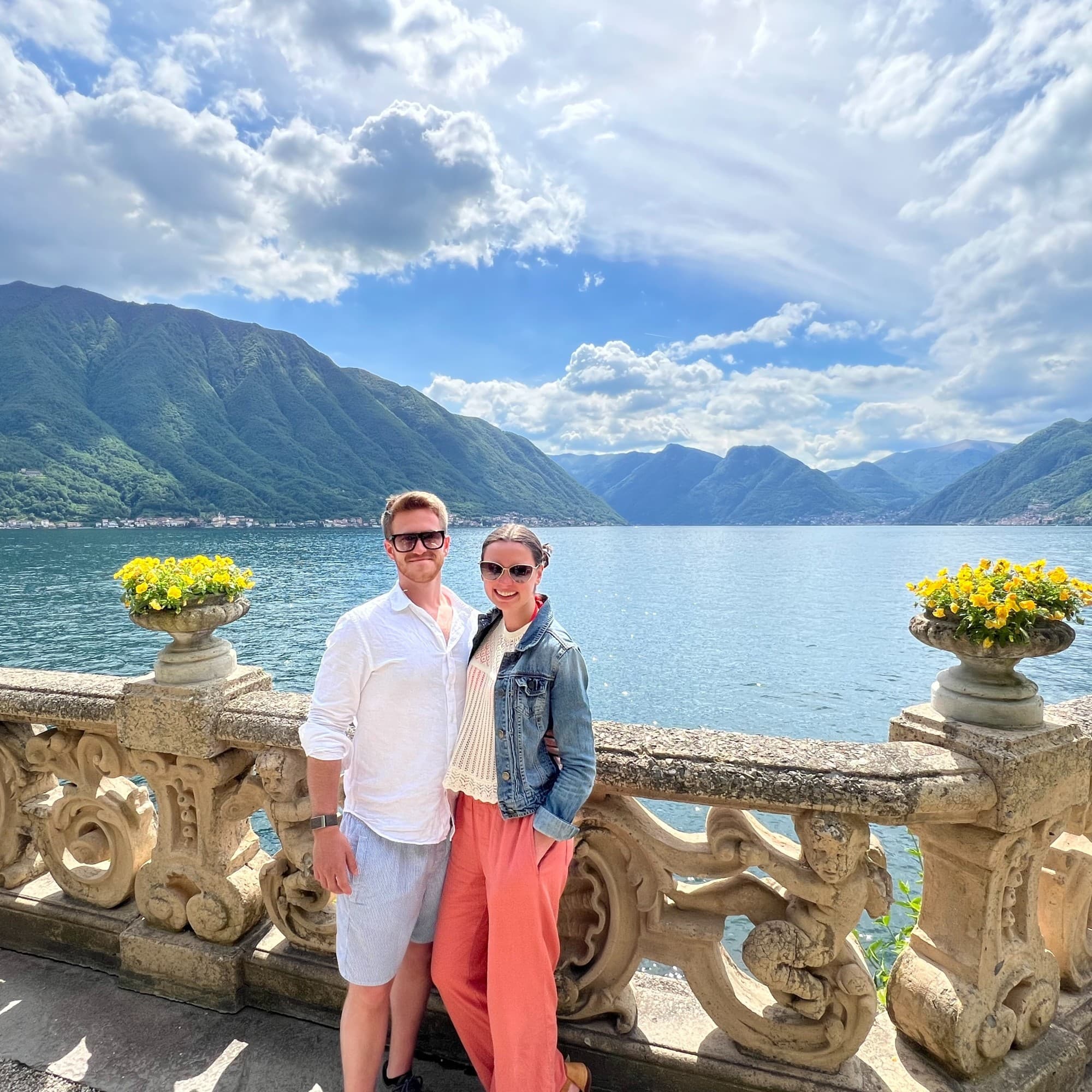 Two people stand by a balustrade with potted flowers, overlooking a scenic lake and mountains.