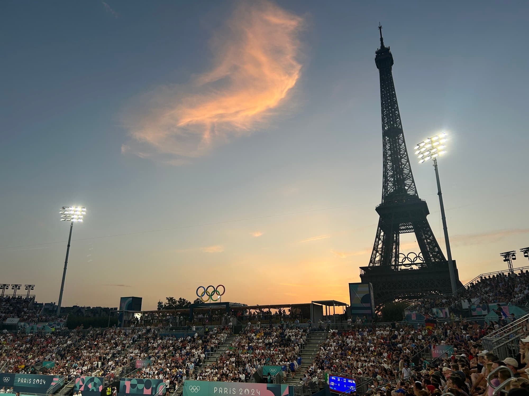 An open-air stadium full of people with a view of a pointed tower in the background during the evening